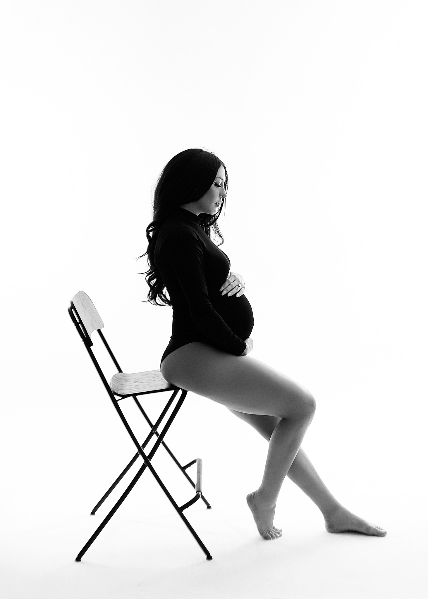 Black and white photo of expecting mom in a black bodysuit sitting in a chair looking down.